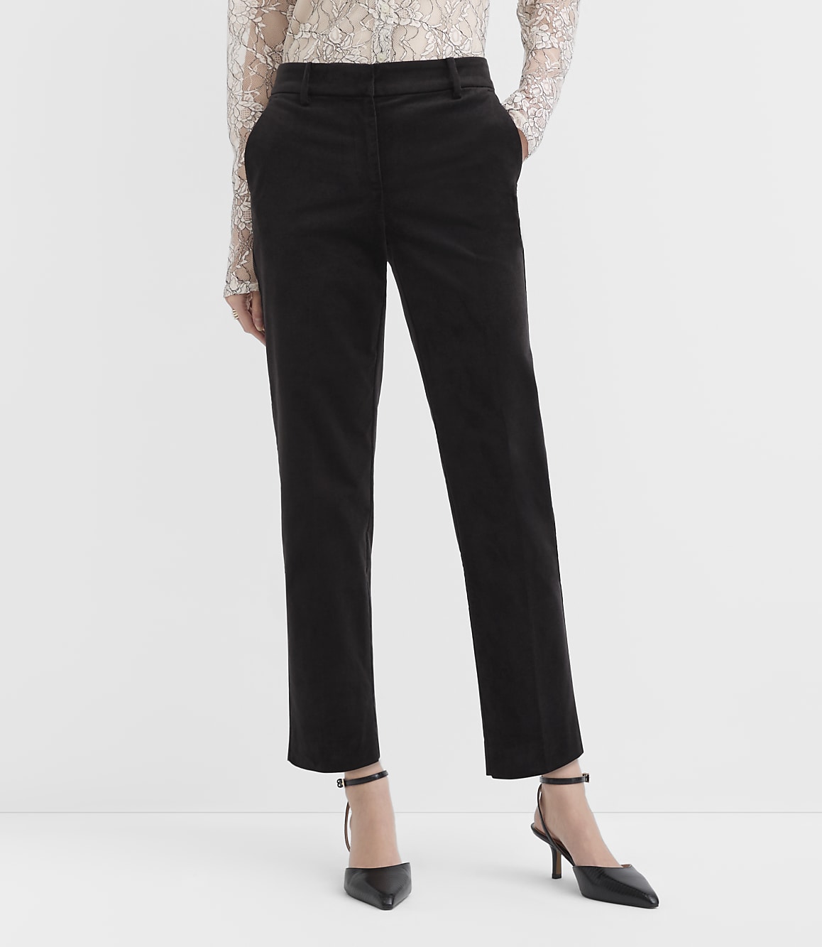A person wearing a lace top, black trousers, and black pointed-toe heels stands against a plain white background.