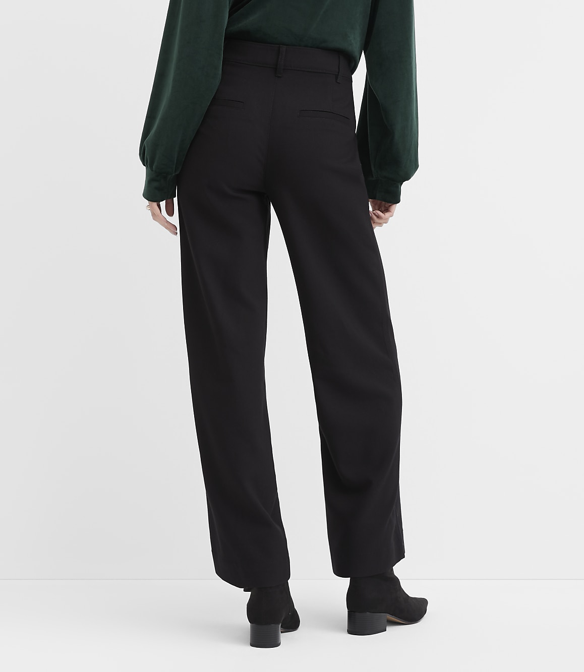 A person wearing a dark green top and black wide-leg trousers stands against a plain white background.