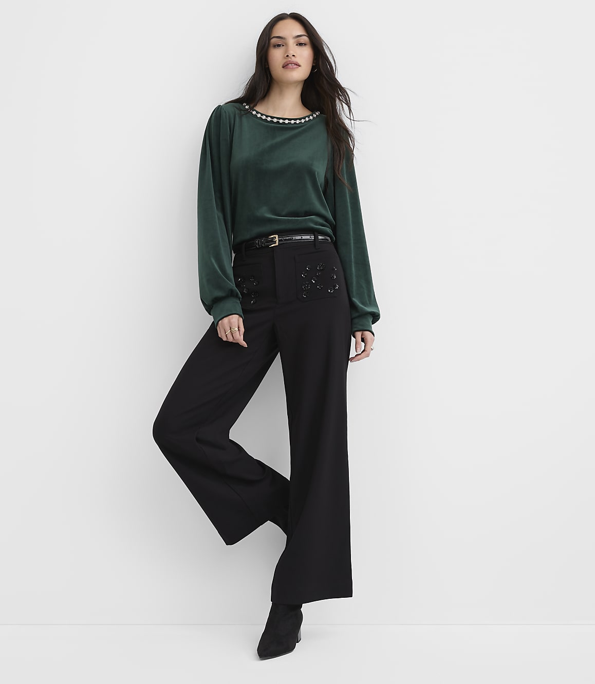 A woman wearing a green long-sleeved top and black wide-leg trousers stands against a plain white background.