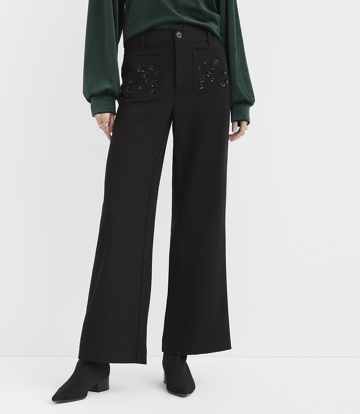 A person wearing a green top, black wide-leg trousers with embellished pockets, and black ankle boots stands against a plain white background.