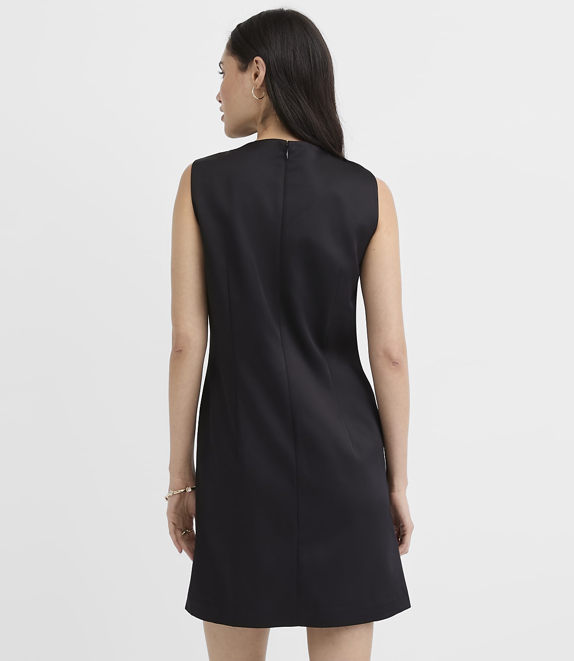 A woman wearing a sleeveless black dress stands with her back to the camera against a plain white background.