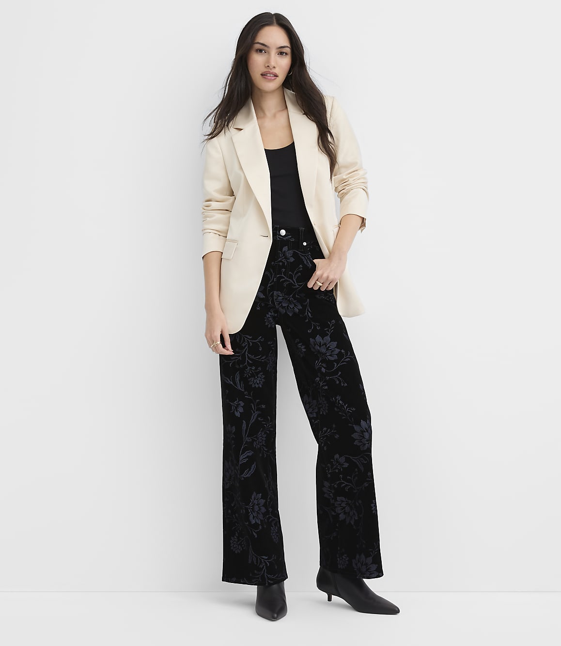 A woman wearing a cream blazer and floral black trousers stands against a plain white background.