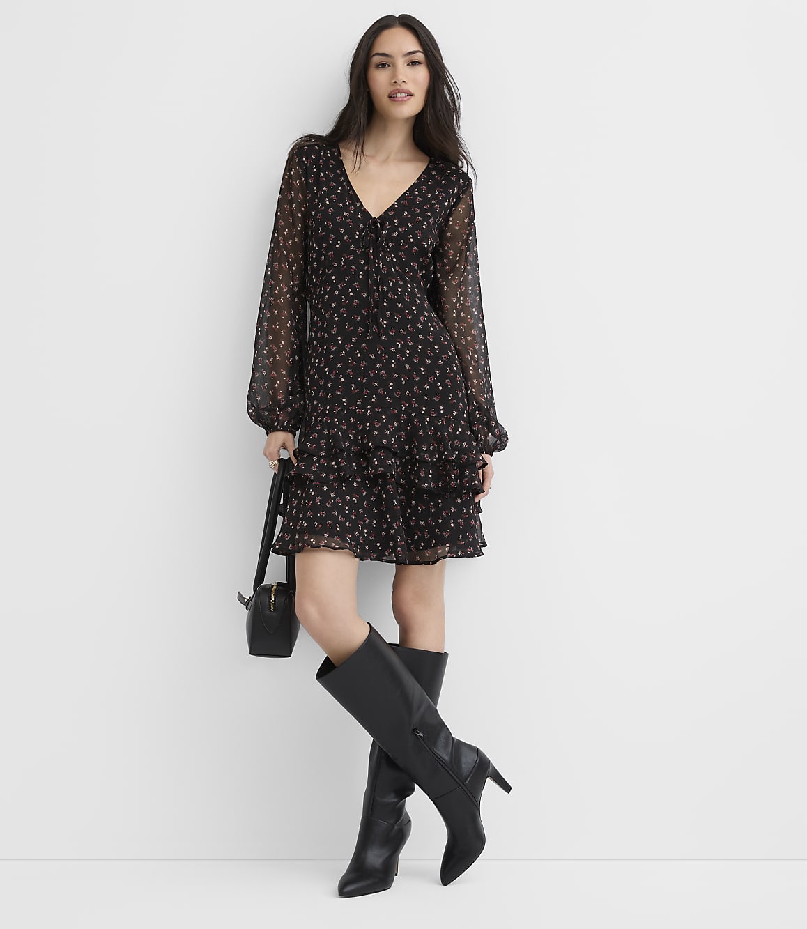 A woman wearing a black floral dress and tall black boots stands against a plain white background.