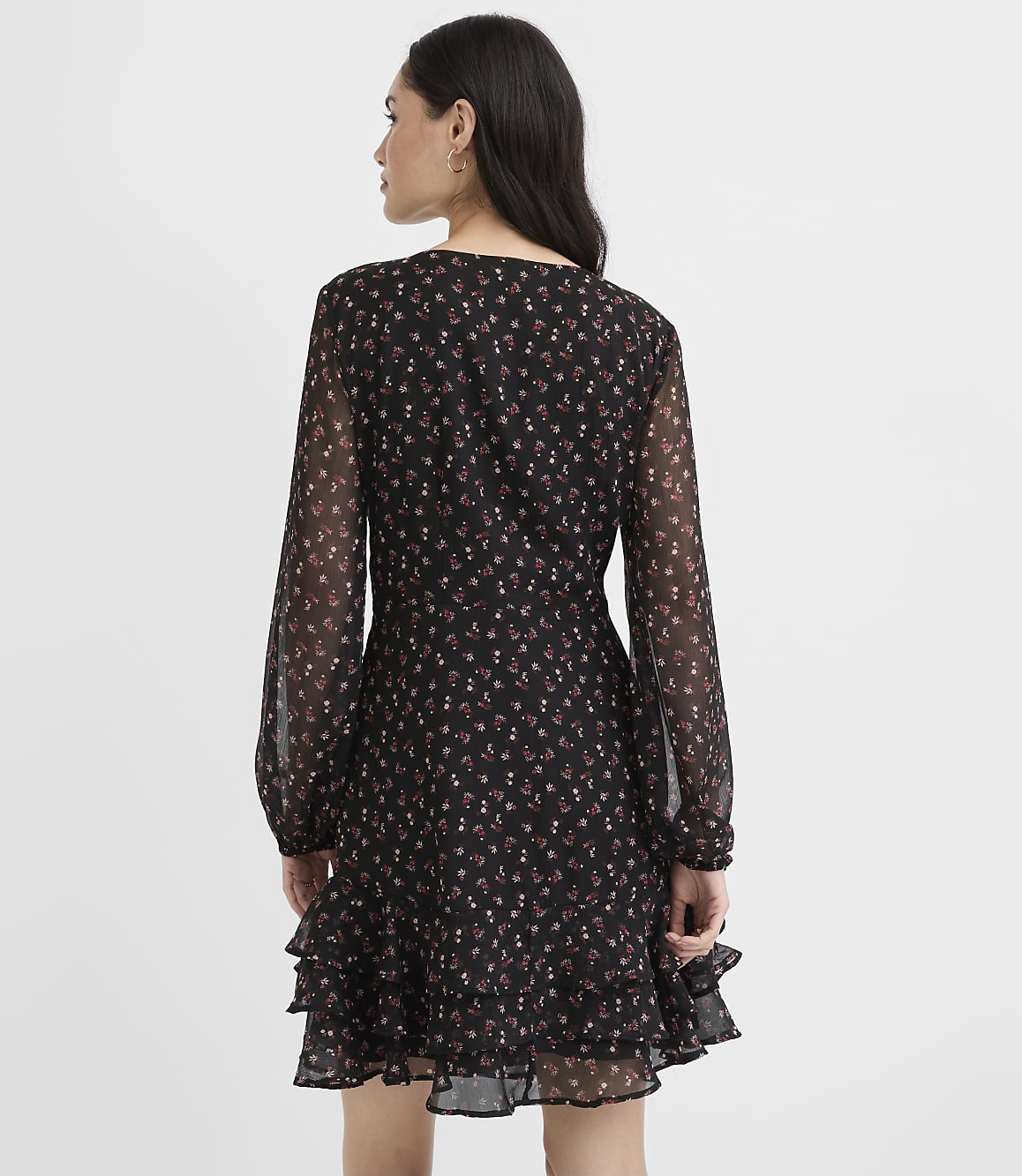A woman stands with her back to the camera wearing a black floral mini dress against a plain white background.