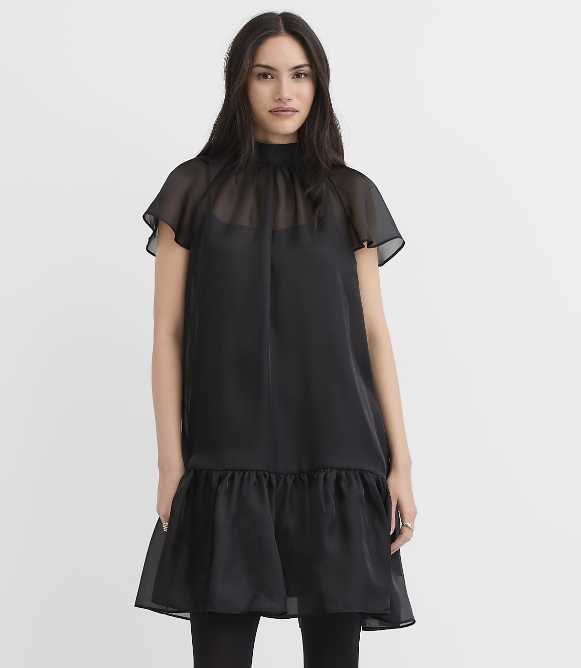 A woman wearing a black dress with sheer sleeves and a ruffled hem stands against a plain white background.