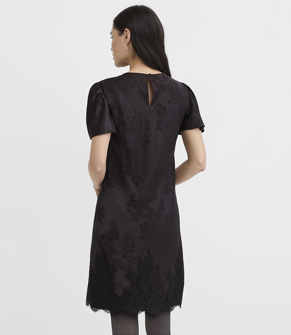A woman stands with her back to the camera wearing a black floral dress against a plain white background.