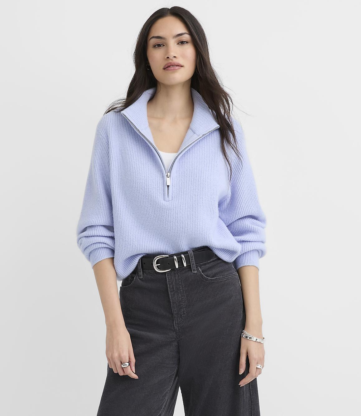 A woman wearing a light blue quarter-zip sweater and dark grey corduroy pants stands against a plain white background.