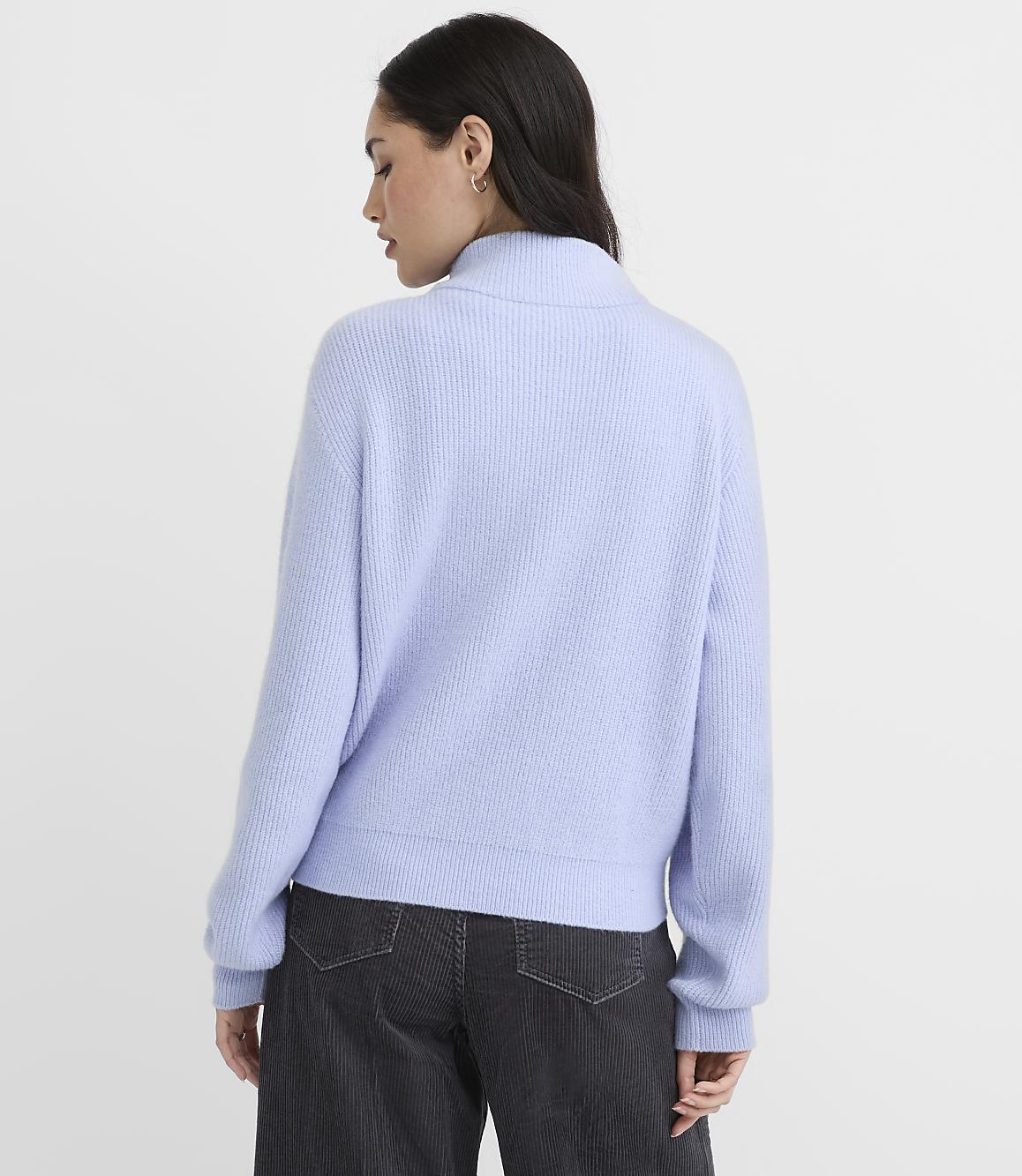 A woman wearing a light purple ribbed sweater and dark grey pants stands against a plain white background.