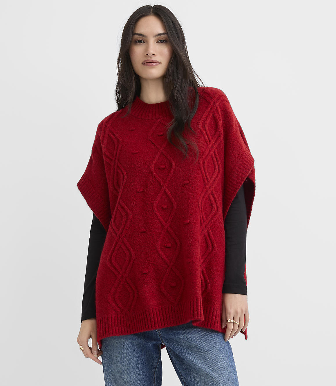 A woman wearing a red cable-knit poncho over a black long-sleeved shirt and blue jeans stands against a plain white background.