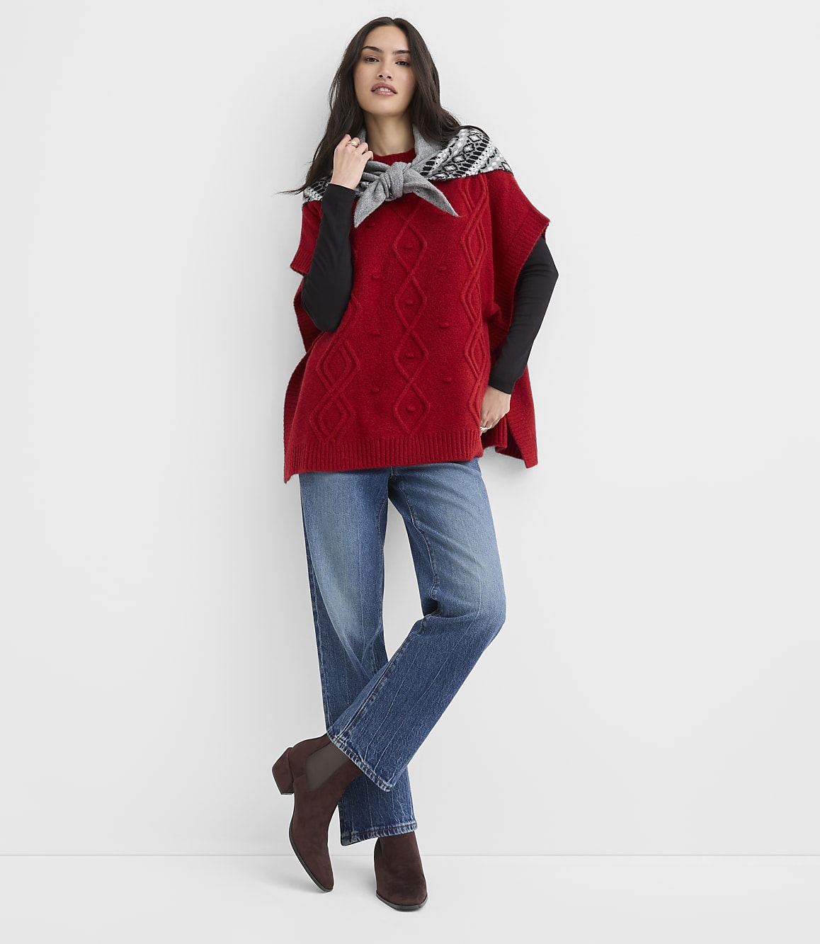 A woman wearing a red cable-knit poncho, black long-sleeved shirt, and blue jeans stands against a plain white background.