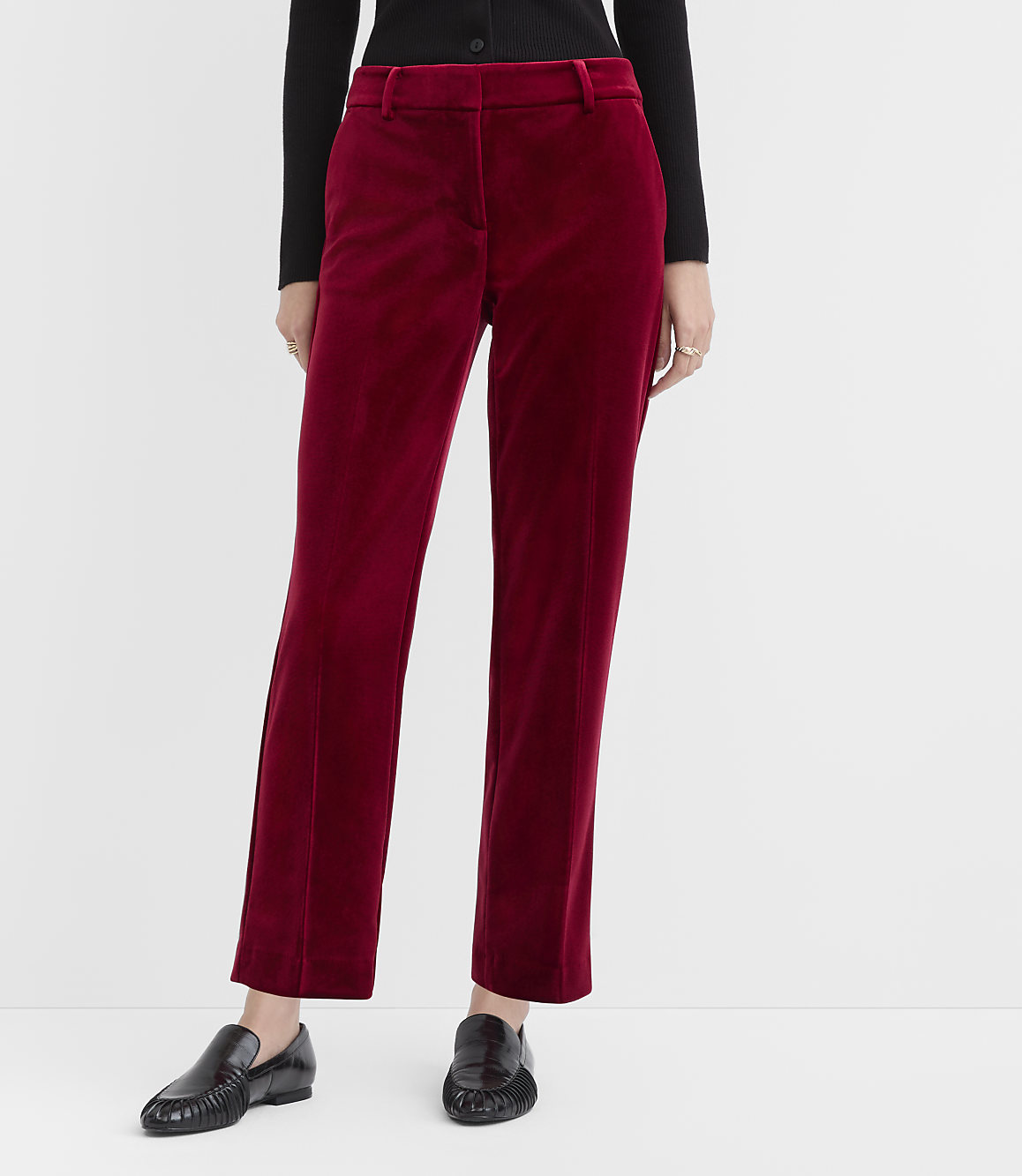 A person wearing a black top, burgundy velvet trousers, and black loafers stands against a plain white background.
