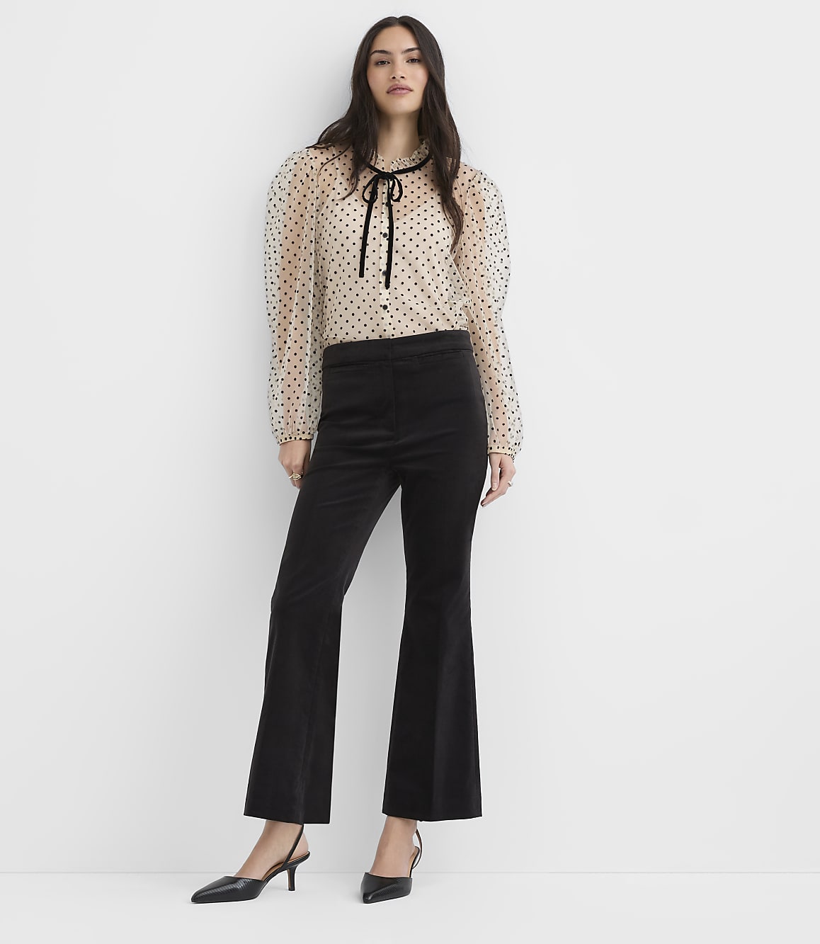 A woman wearing a polka-dot blouse and black flared trousers stands against a plain white background.