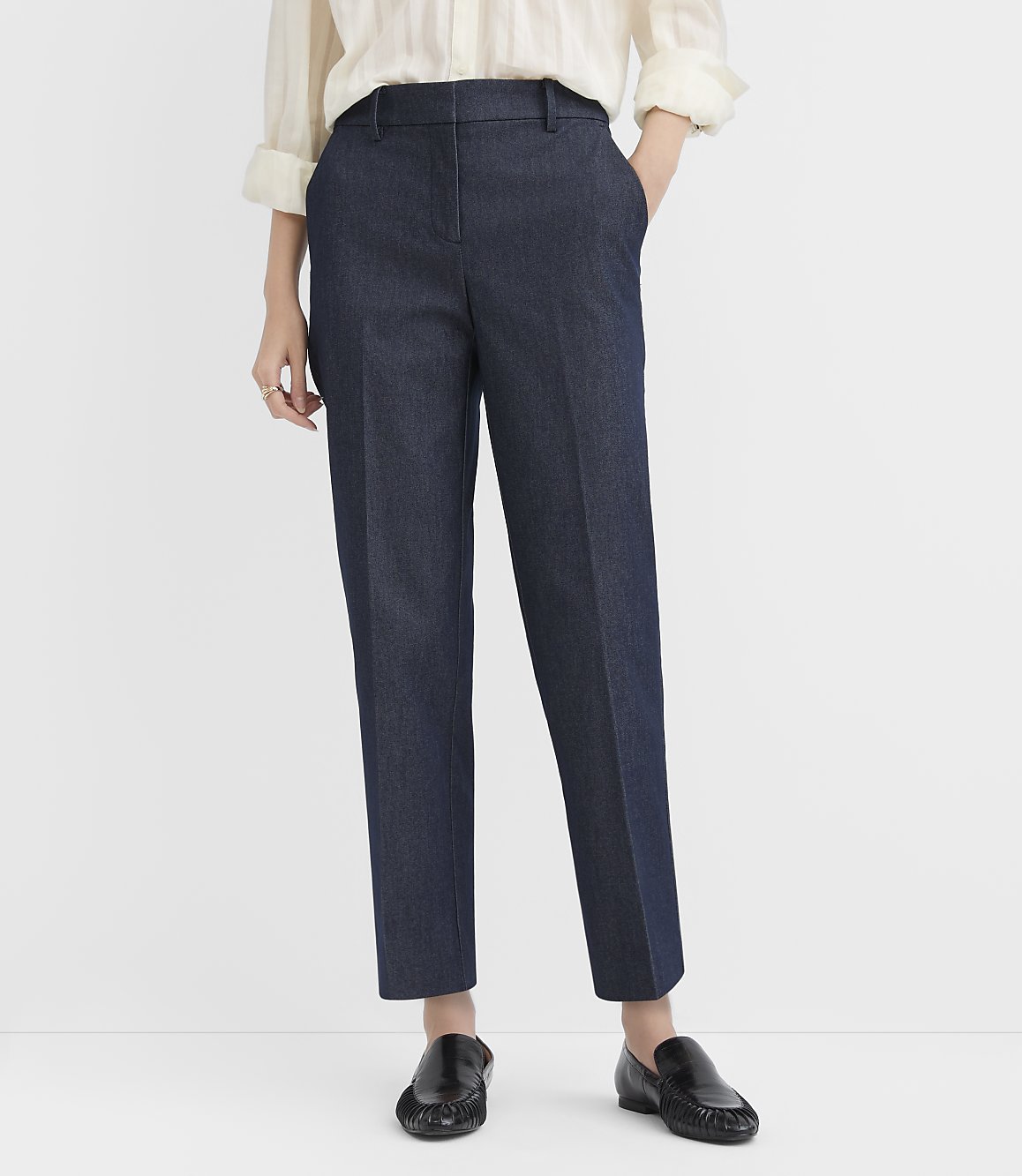A person wearing a white blouse, navy blue trousers, and black loafers stands against a plain white background.