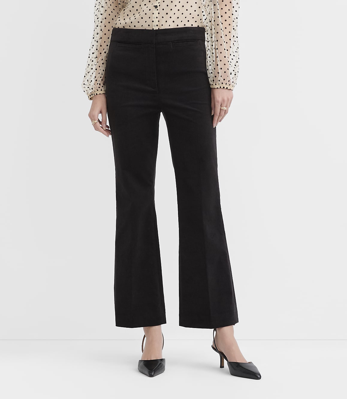 A person wearing a polka-dot blouse, black flared trousers, and black pointed-toe heels stands against a plain white background.