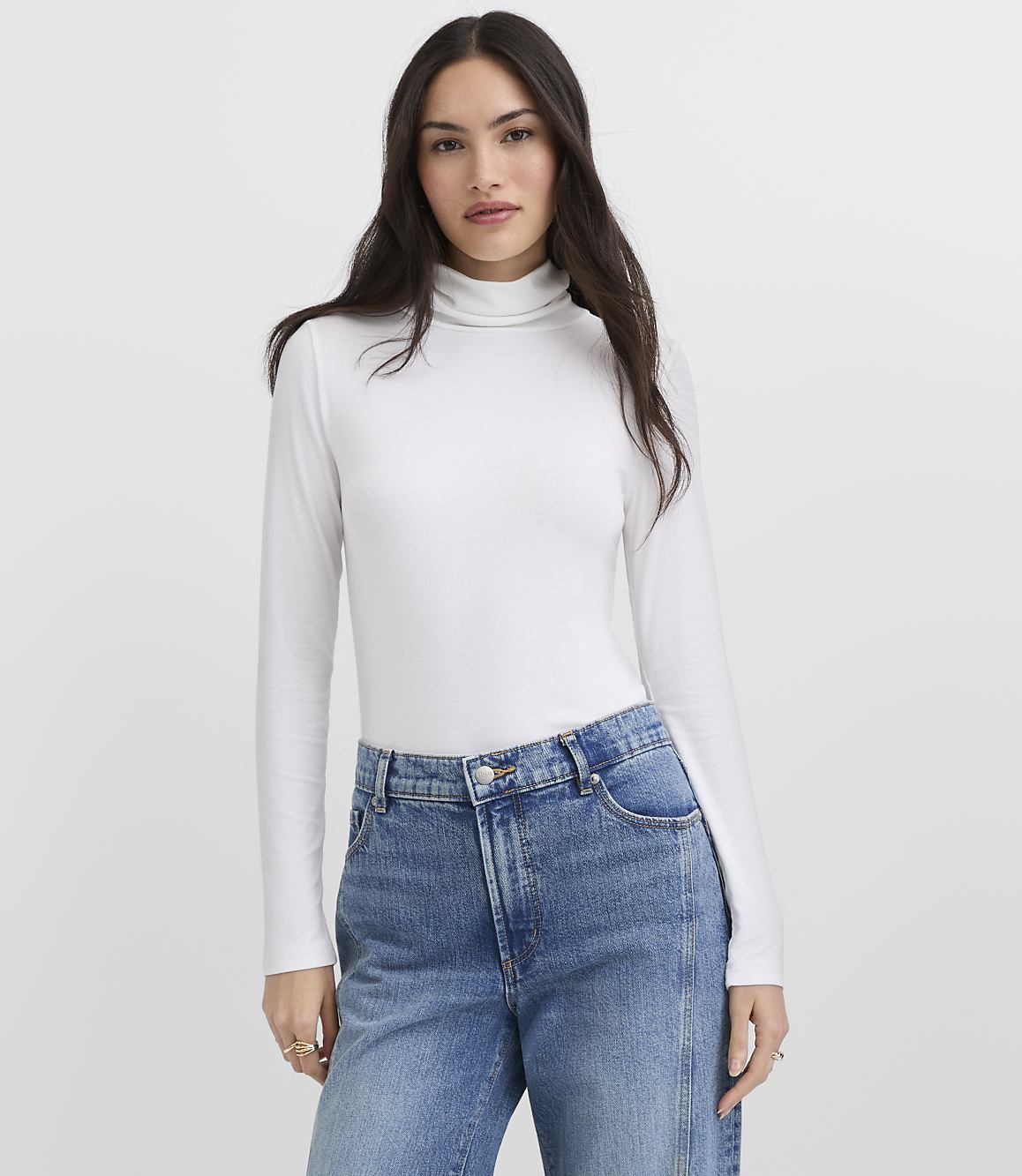 A woman wearing a white turtleneck and blue jeans stands against a plain light gray background.