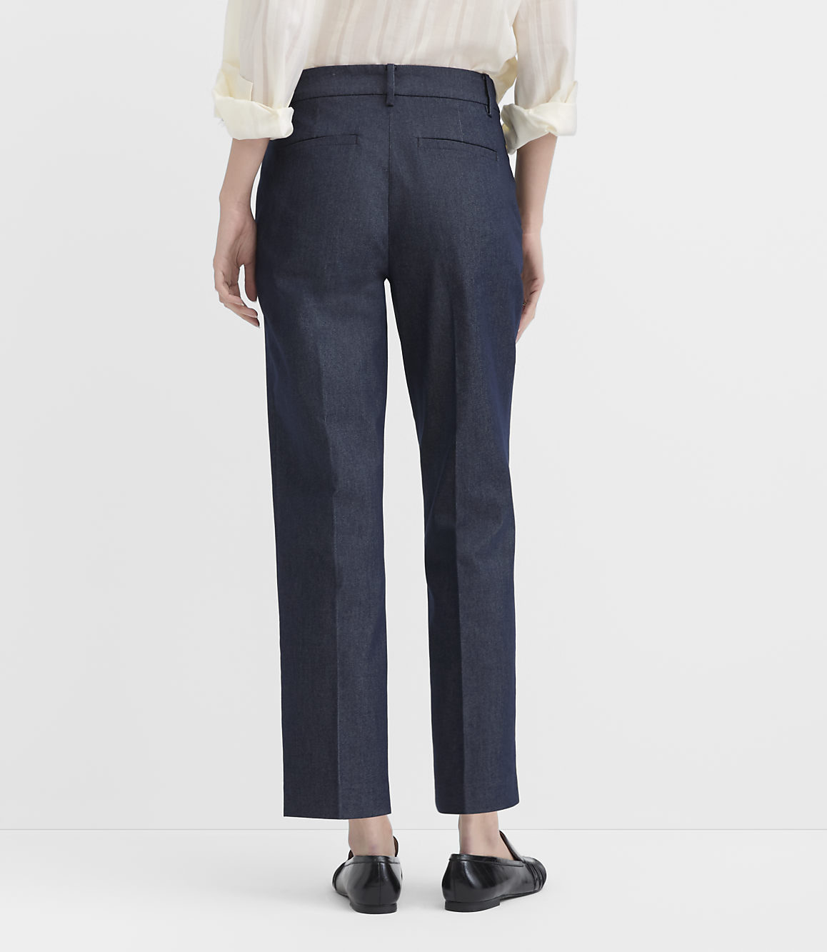 A person stands with their back to the camera wearing a cream blouse, dark blue trousers, and black flats against a plain white background.