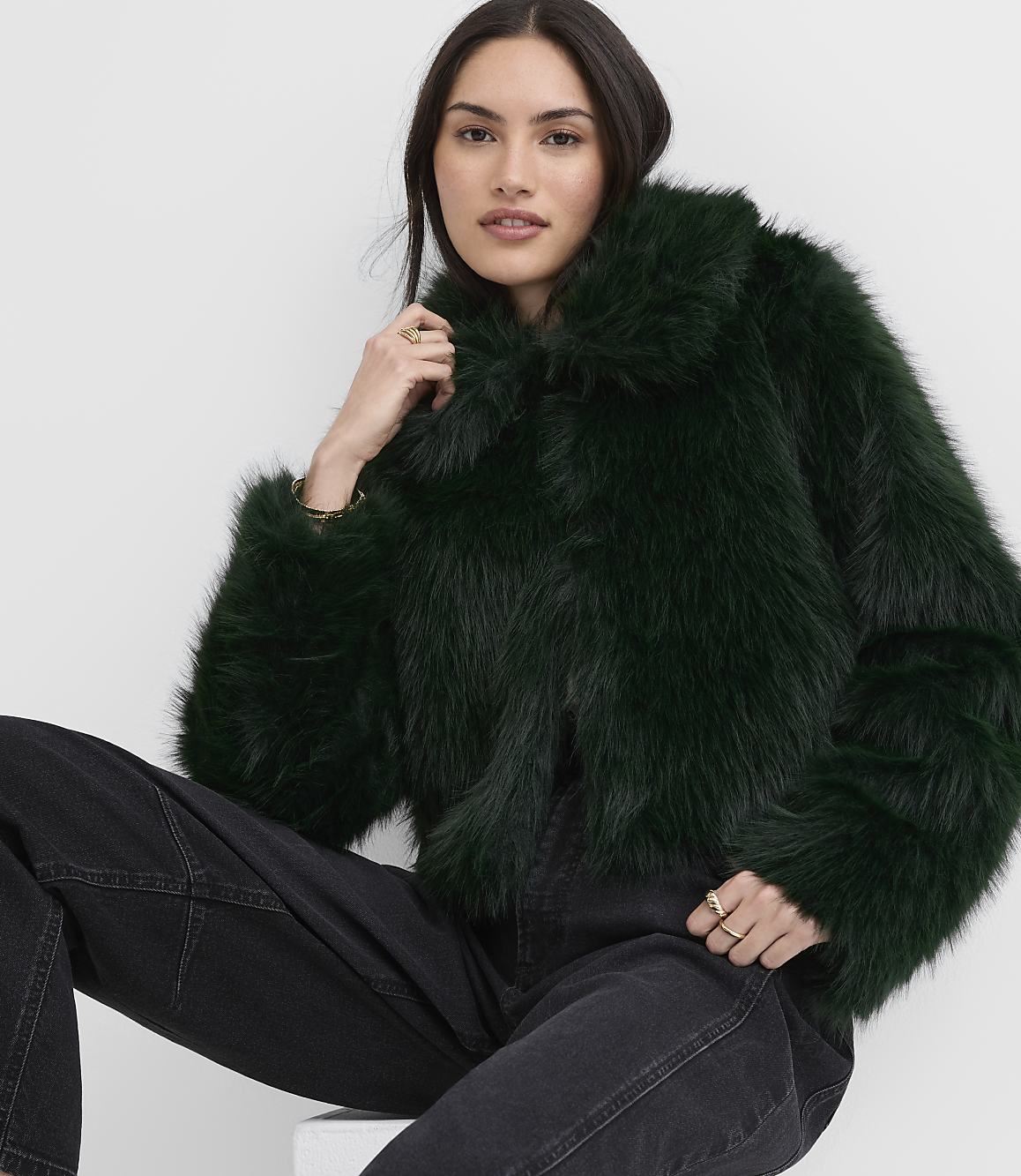 A woman wearing a dark green faux fur jacket and black jeans poses against a plain white background.