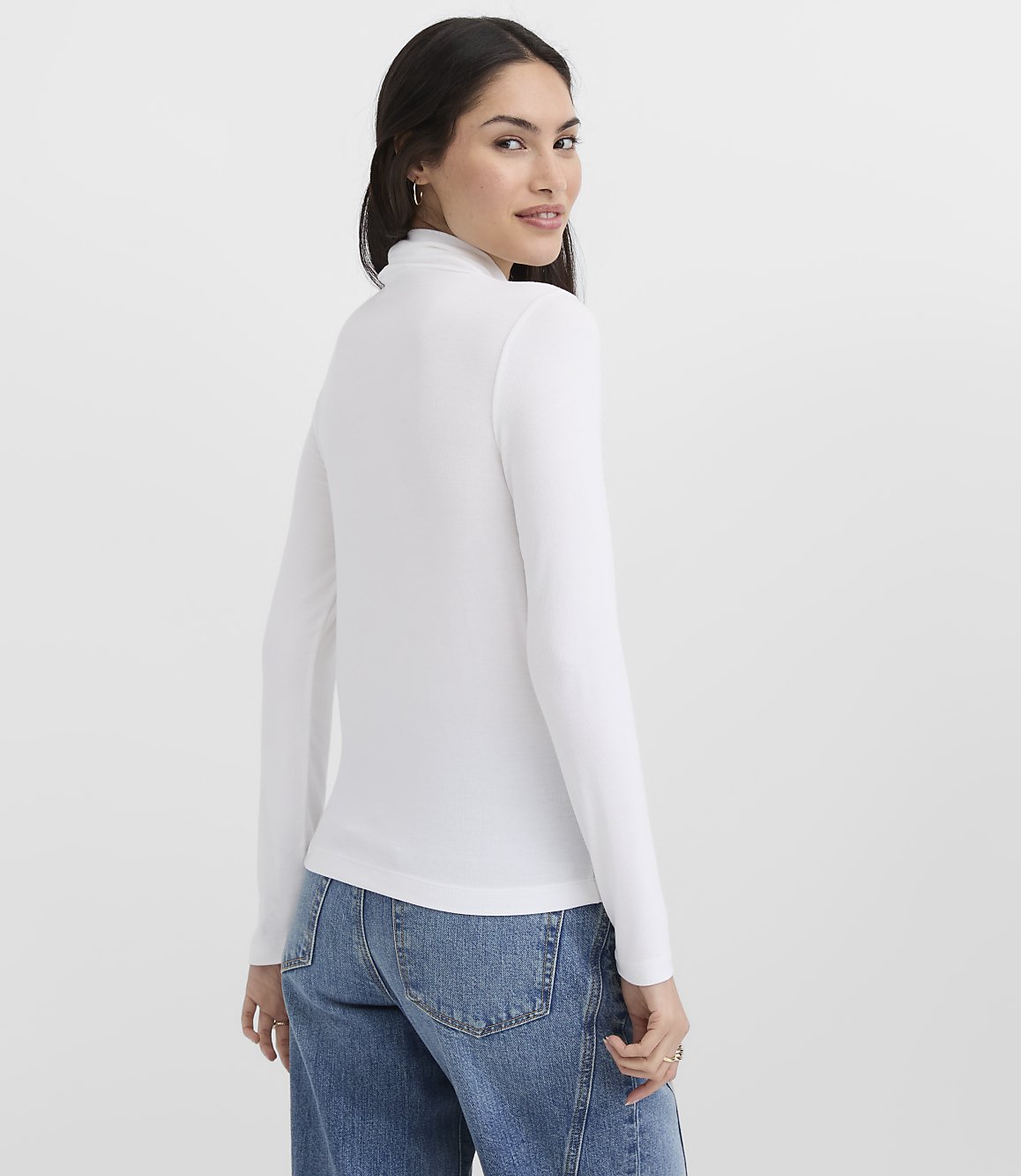 A woman wearing a white long-sleeved turtleneck and blue jeans stands against a plain white background.