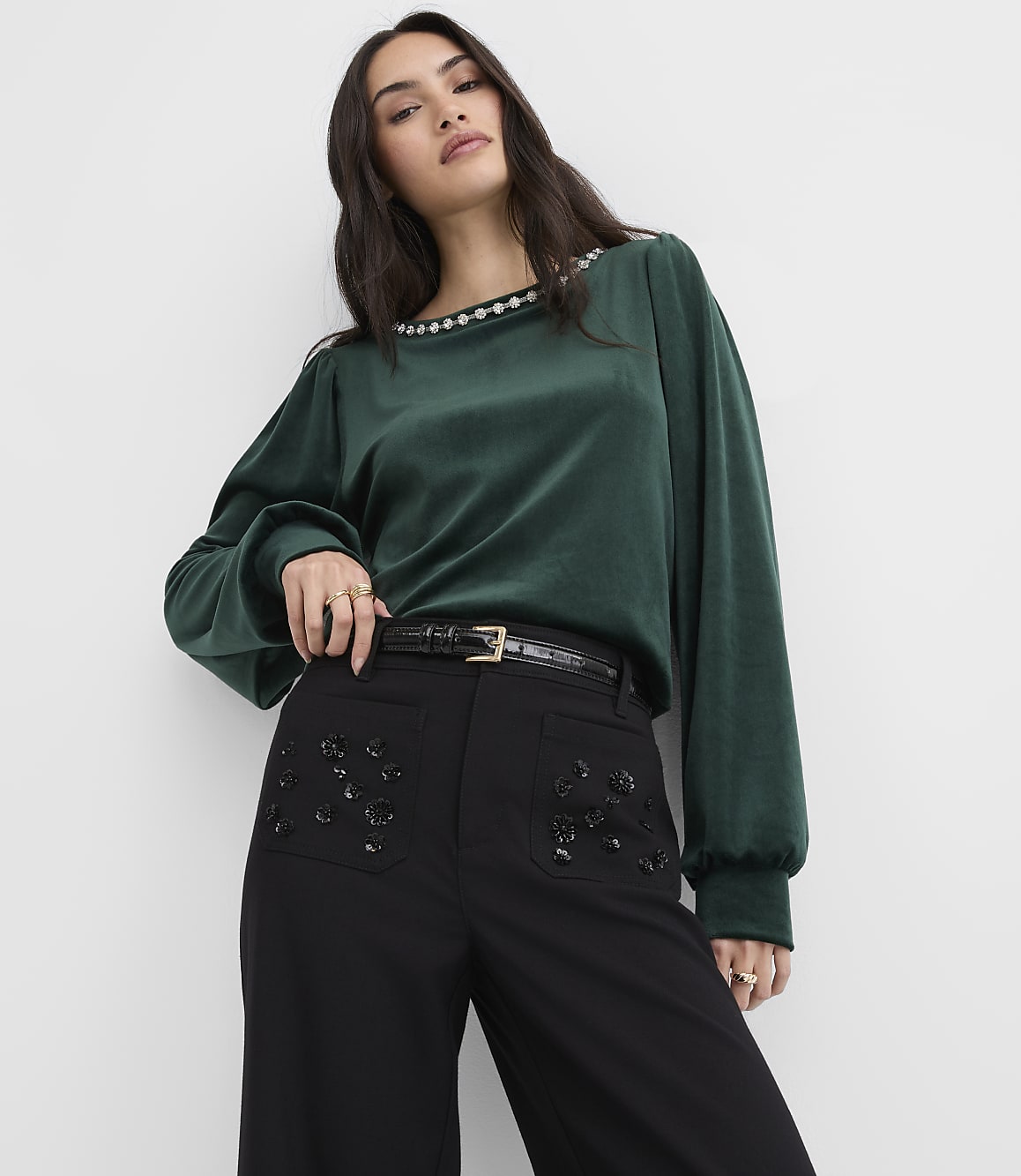 A woman wearing a dark green blouse and black embellished trousers stands against a plain white background.