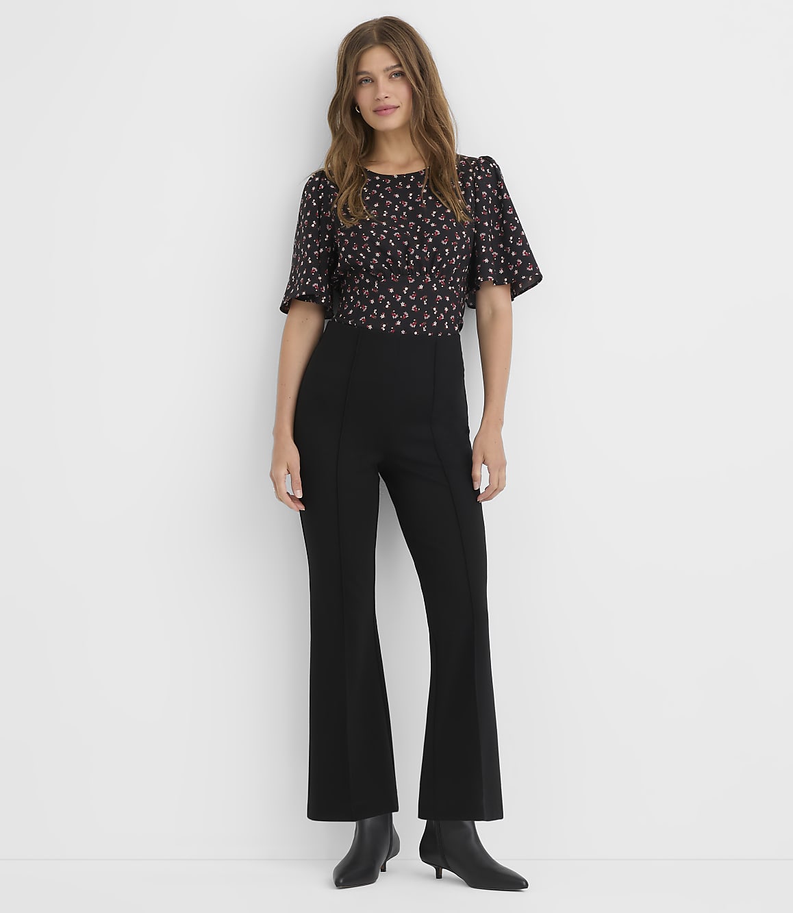 A woman in a floral top and black flared pants stands against a plain white background.