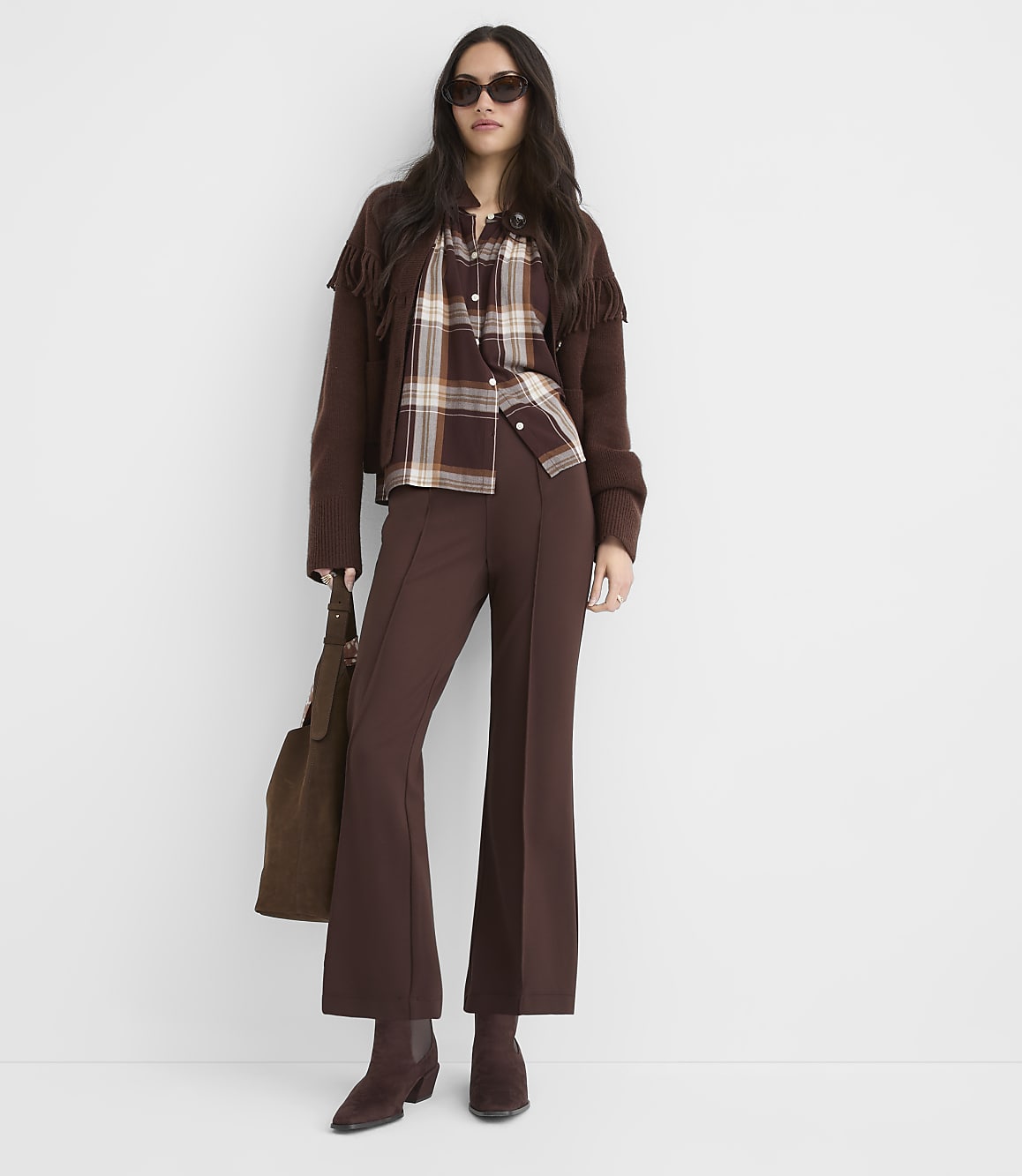 A woman in a brown plaid shirt, cropped jacket with fringe, and flared pants stands against a plain white background, holding a brown suede bag.