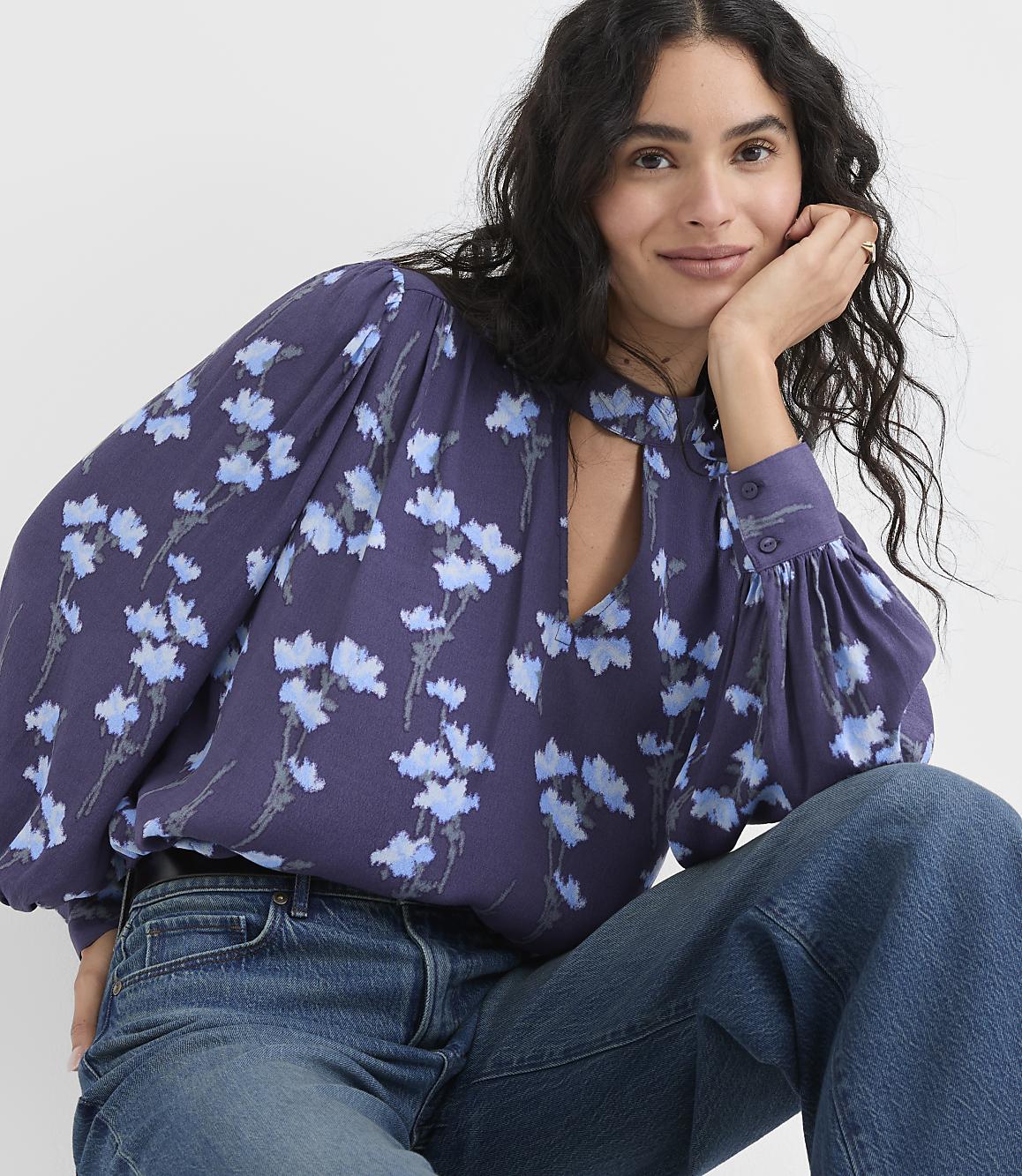 A woman wearing a purple floral blouse and blue jeans poses against a plain white background.