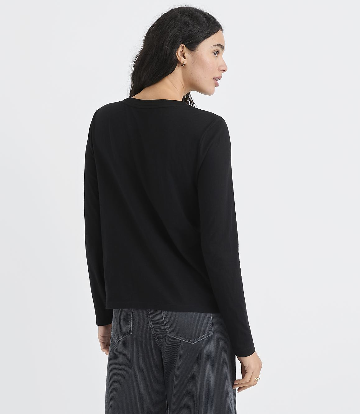 A woman wearing a black long-sleeved shirt and dark grey trousers stands against a plain white background.