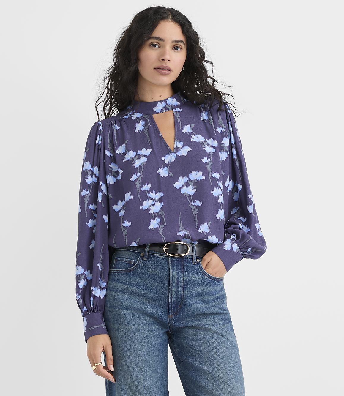 A woman wearing a purple floral blouse and blue jeans stands against a plain white background.