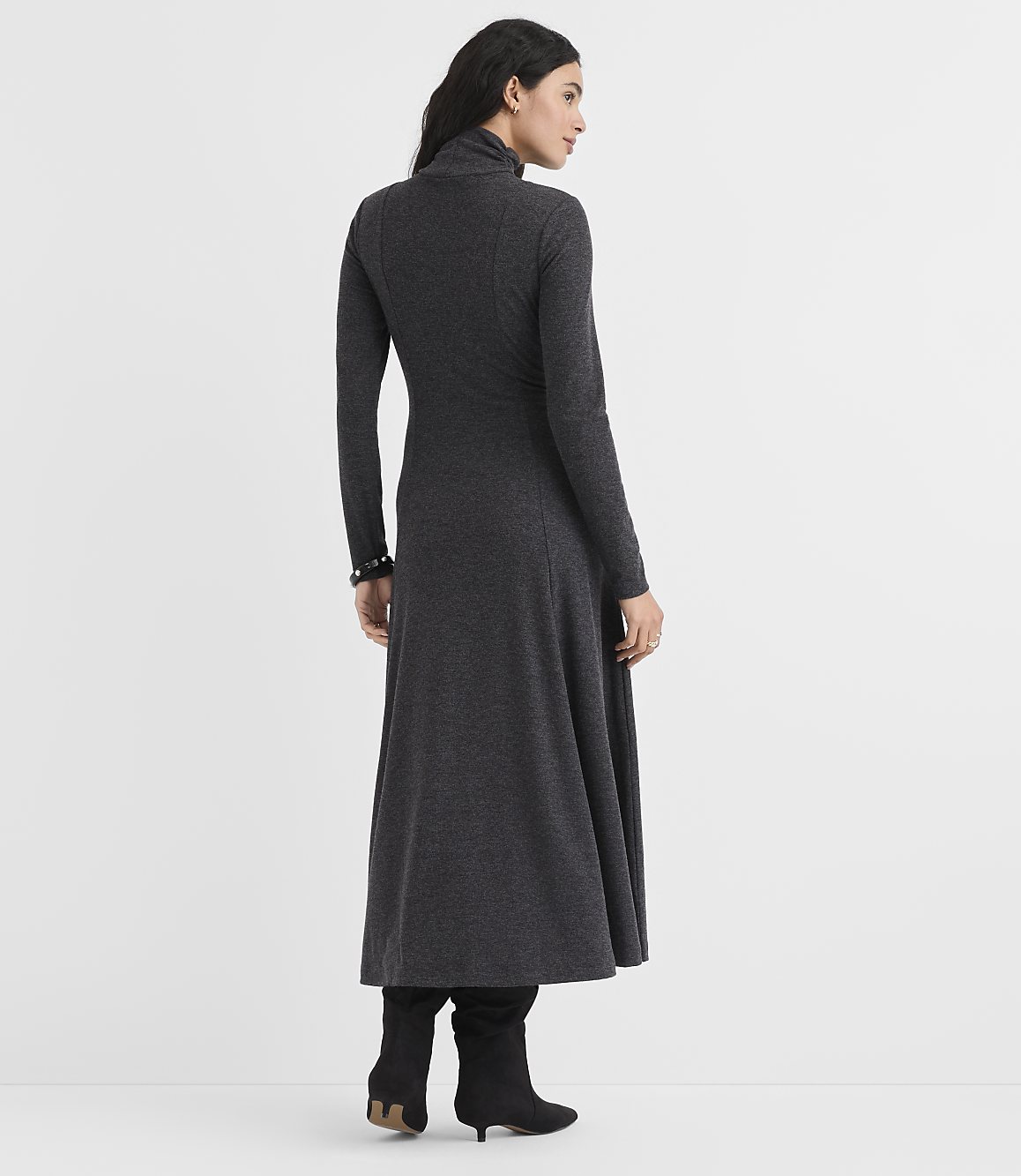 A woman wearing a long-sleeved, charcoal grey maxi dress and black boots stands against a plain white background.