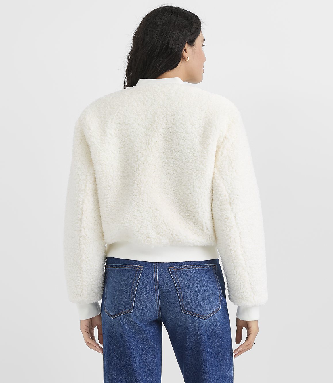 A woman wearing a white fuzzy jacket and blue jeans stands with her back to the camera against a plain light gray background.