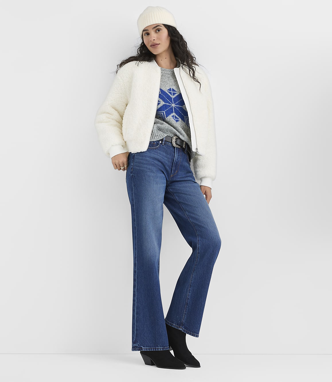 A woman wearing a white beanie, white fuzzy jacket, patterned sweater, and blue flared jeans stands against a plain white background.