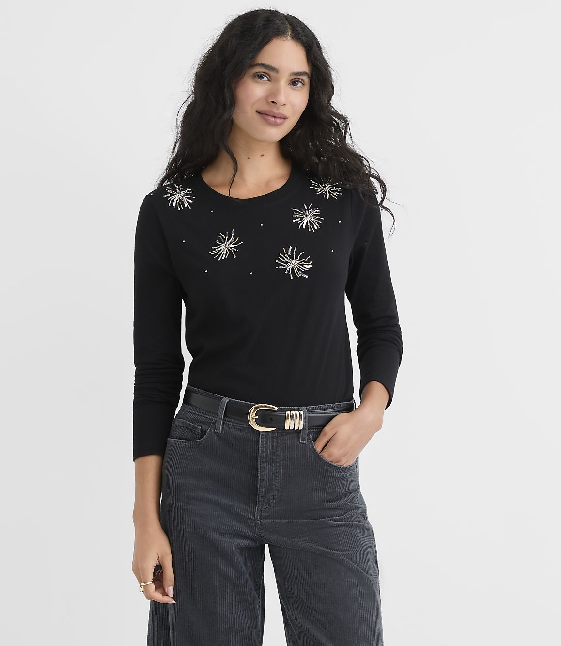 A woman wearing a black sequined top and dark grey corduroy pants stands against a plain white background.