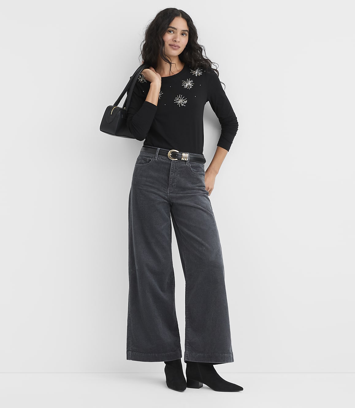 A woman wearing a black sequined top, wide-leg grey trousers, and black boots stands against a plain white background.