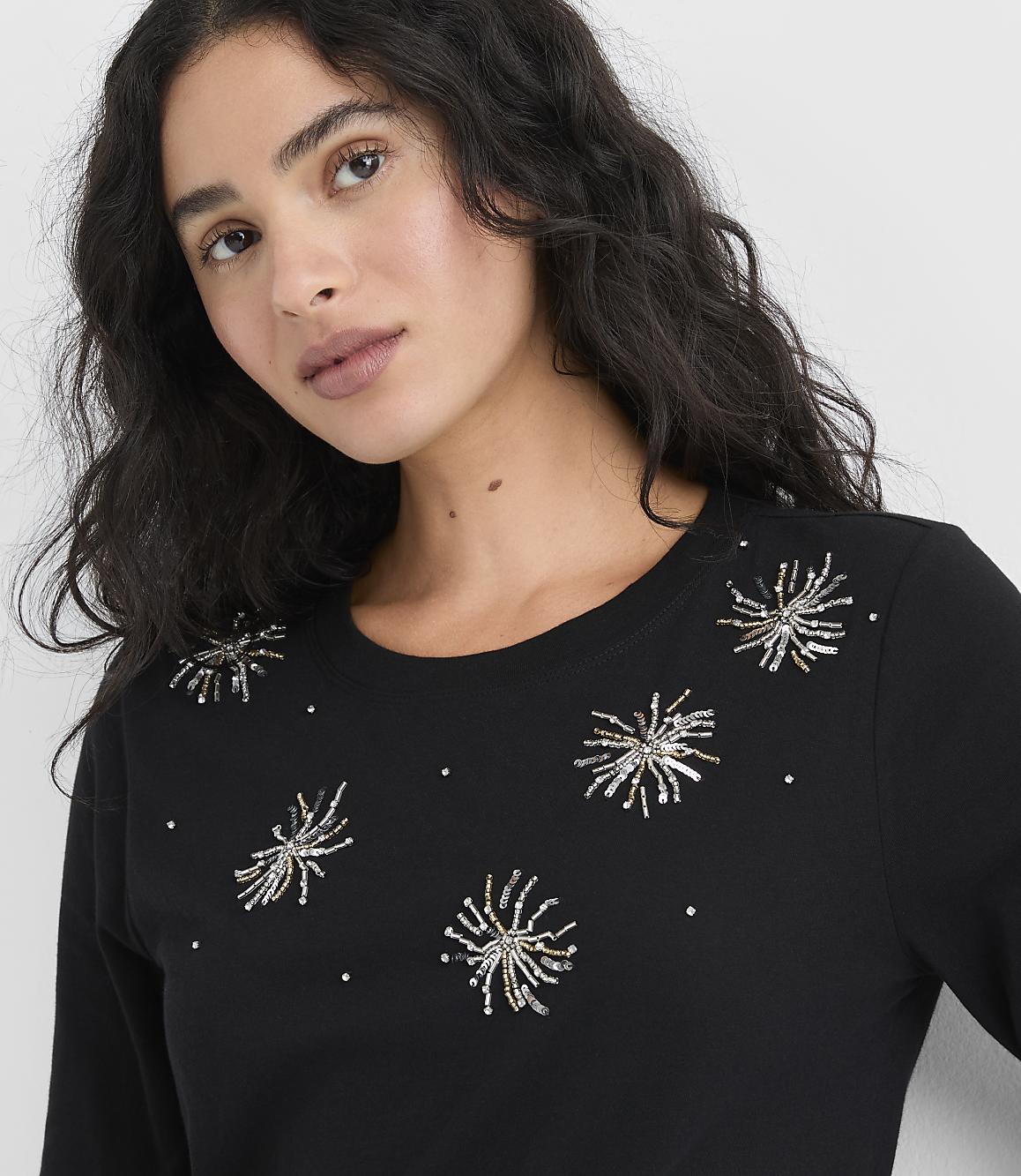 A woman with long, wavy dark hair wearing a black shirt with silver and gold sequin starbursts poses against a plain white background.