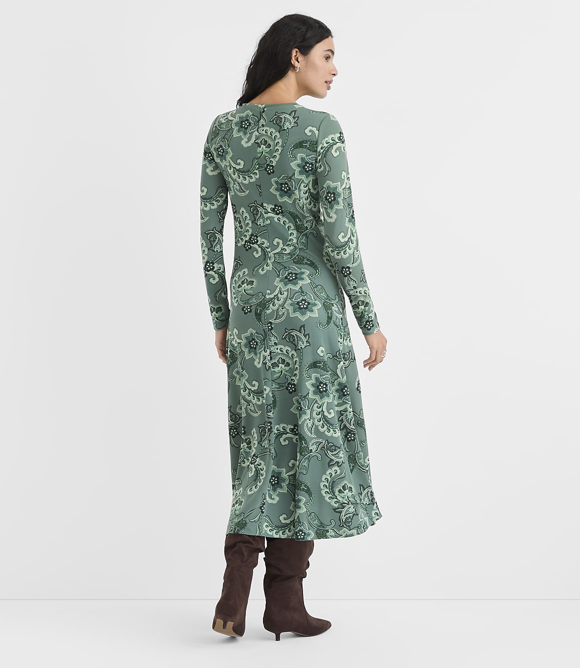 A woman wearing a green paisley print maxi dress and brown boots stands against a plain white background.