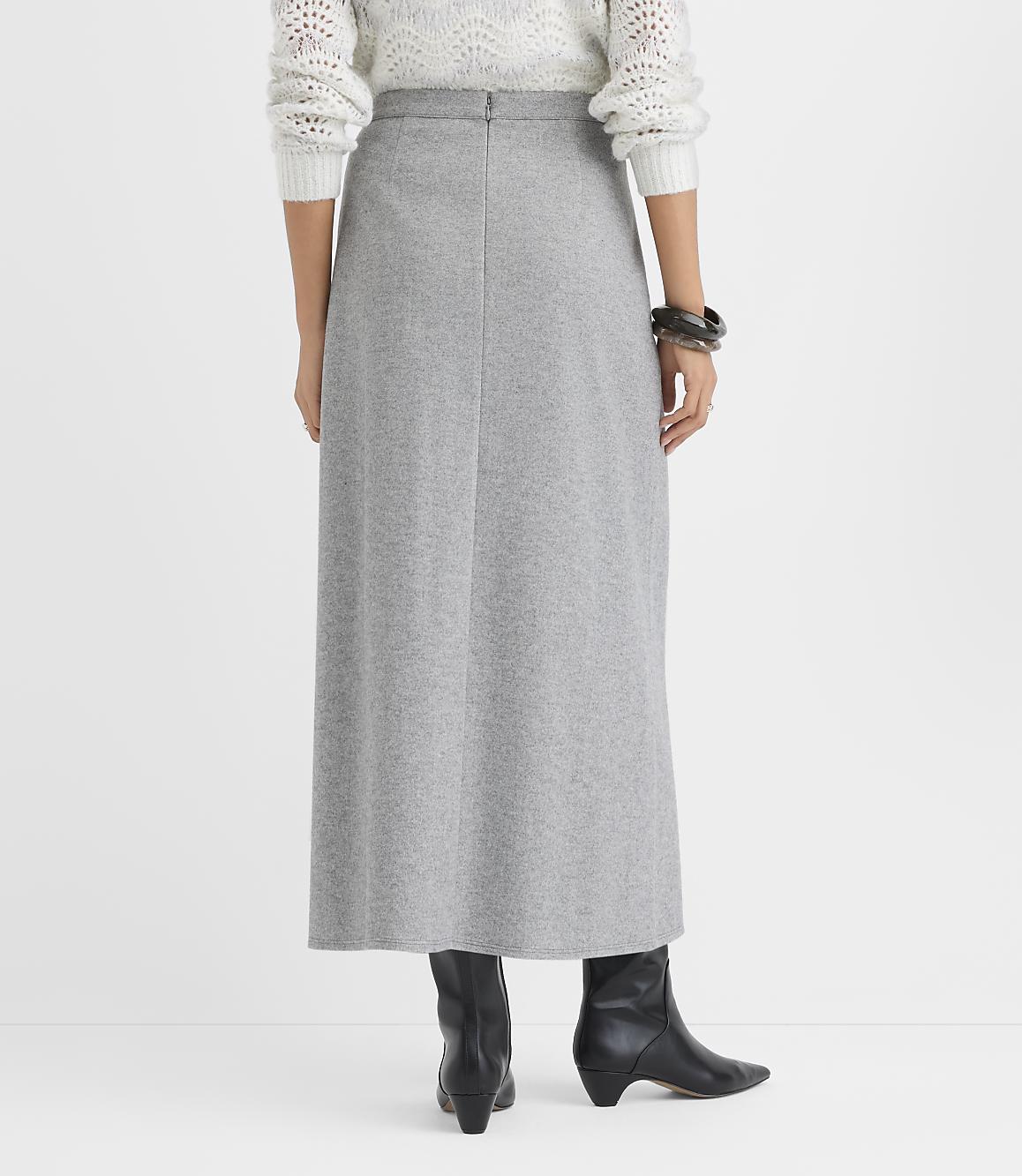 A person wearing a white knit sweater, a long grey skirt, and black ankle boots stands against a plain white background.