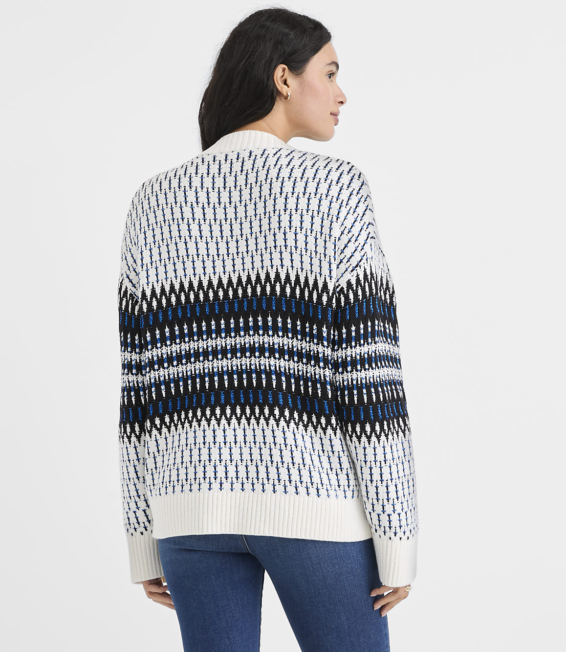 A woman wearing a patterned white, blue, and black sweater and blue jeans stands against a plain white background.