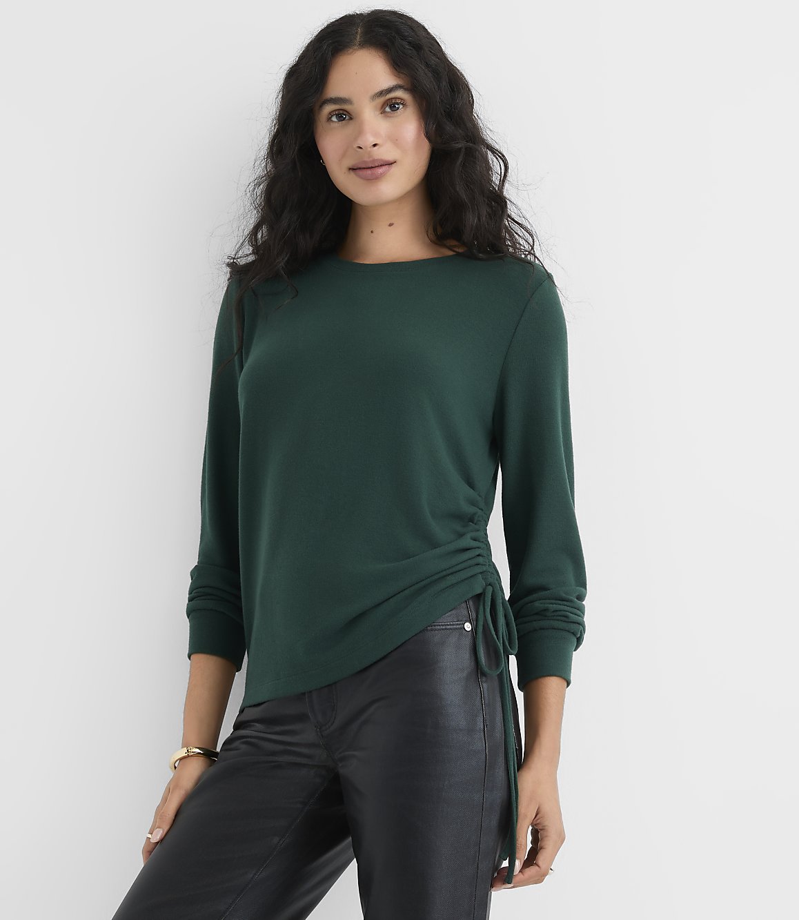 A woman wearing a dark green long-sleeved top and black leather pants stands against a plain white background.