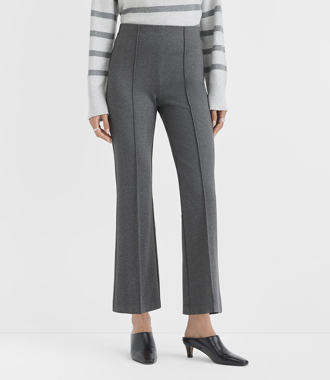 A person wearing a striped sweater, charcoal grey trousers, and black pointed-toe mules stands against a plain white background.