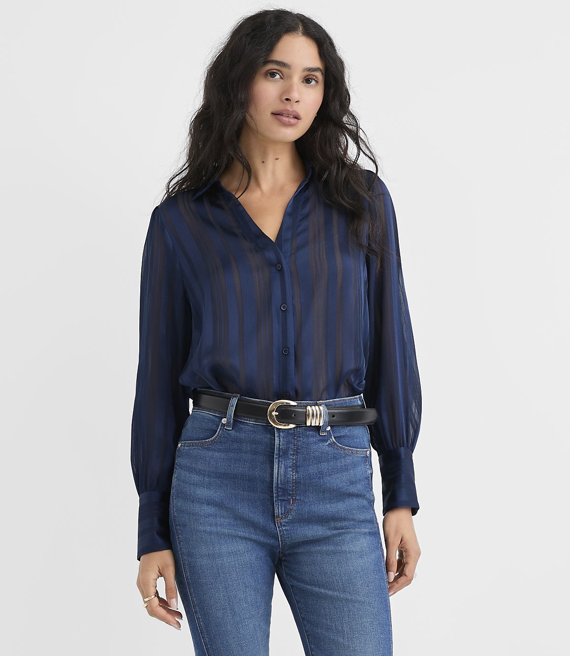 A woman wearing a navy blue striped blouse and blue jeans stands against a plain white background.