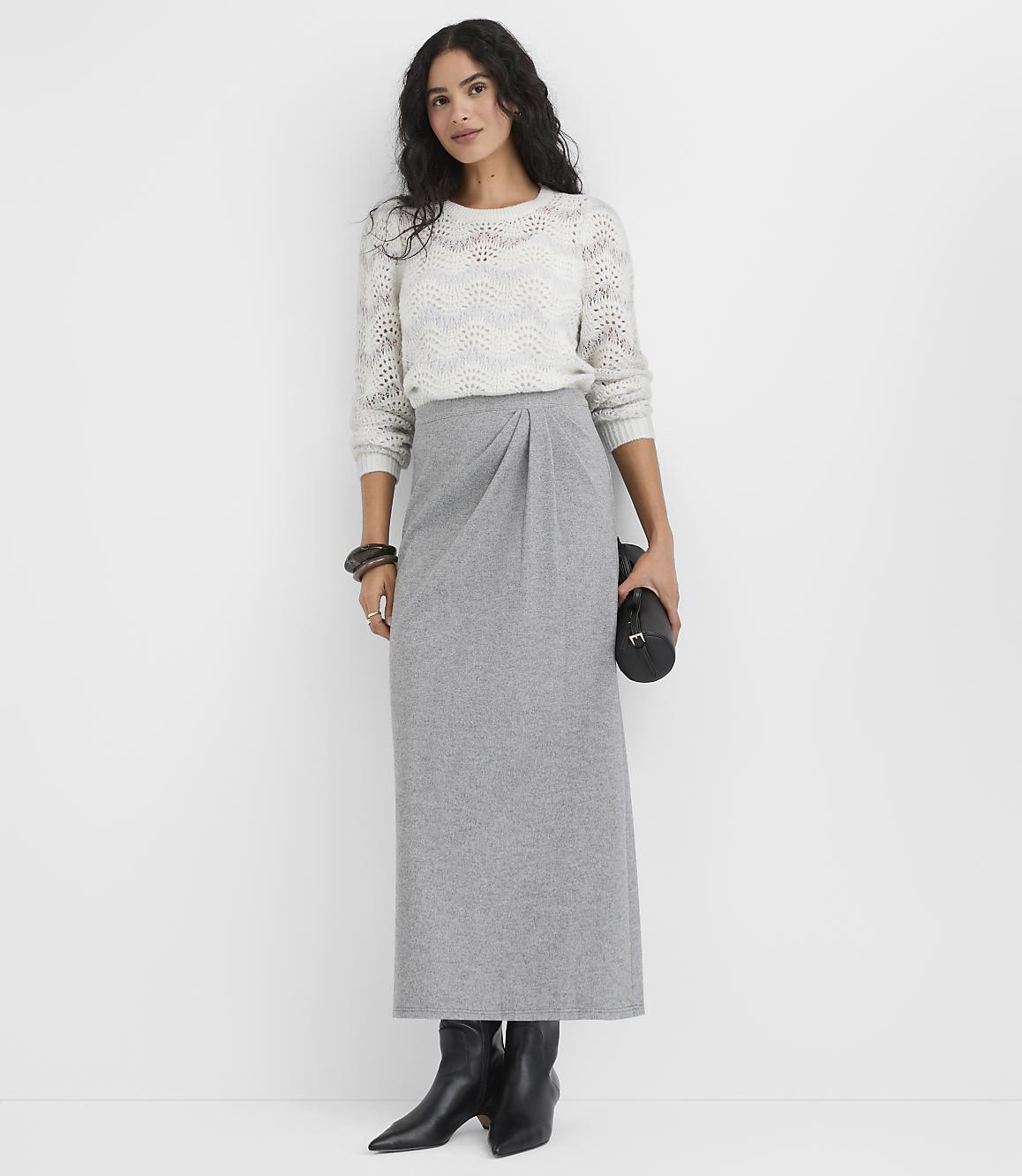 A woman wearing a white knit sweater and a long grey skirt stands against a plain white background.
