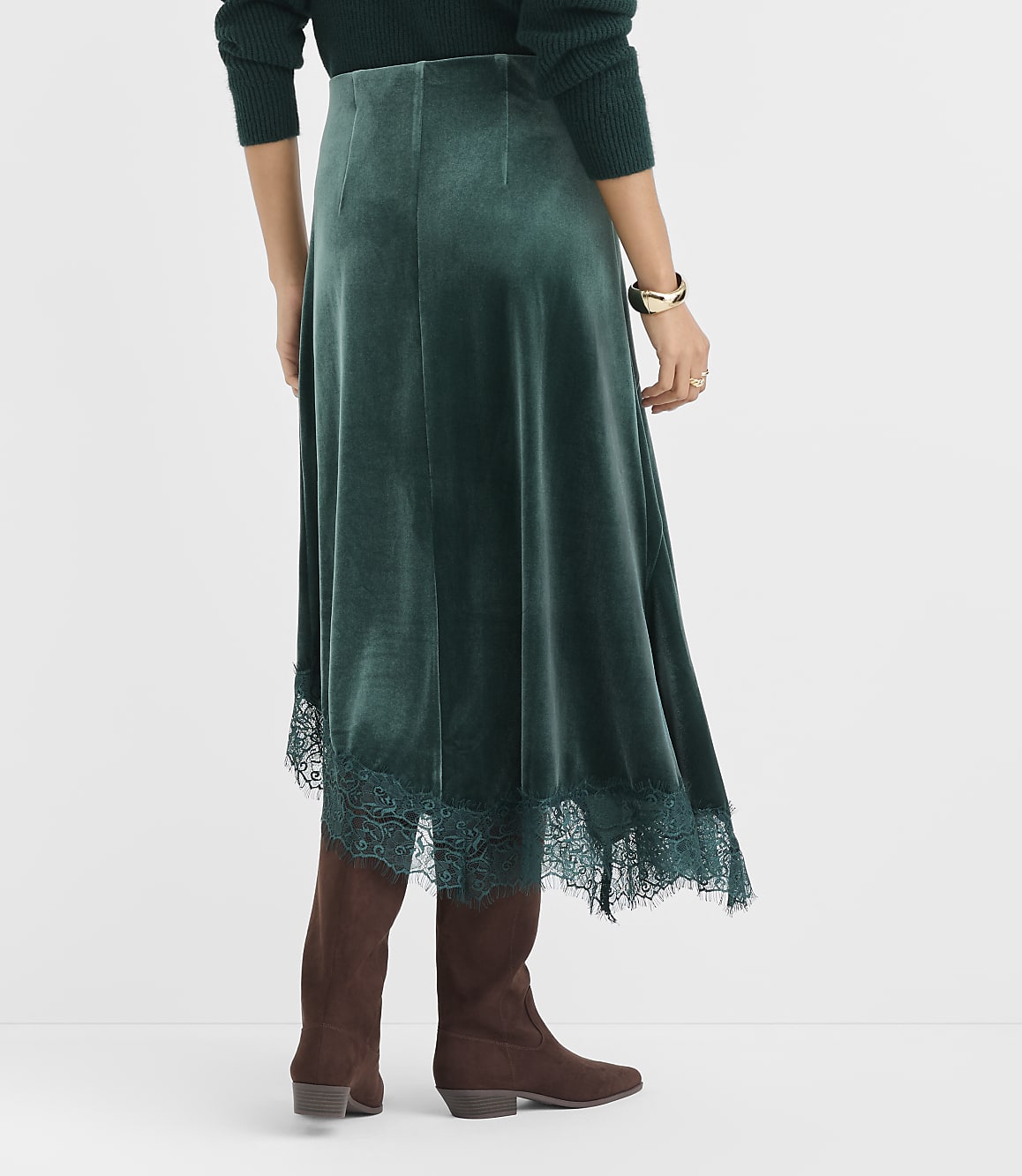 A person wearing a dark green velvet skirt with lace trim and brown boots stands against a plain white background.