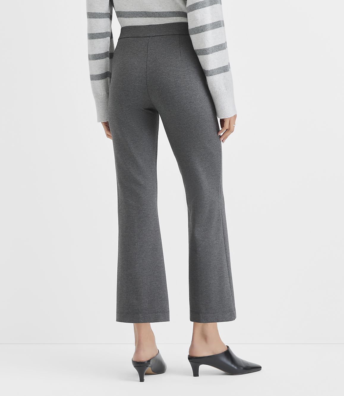 A person wearing a grey striped sweater, charcoal grey cropped trousers, and black mules stands against a plain white background.