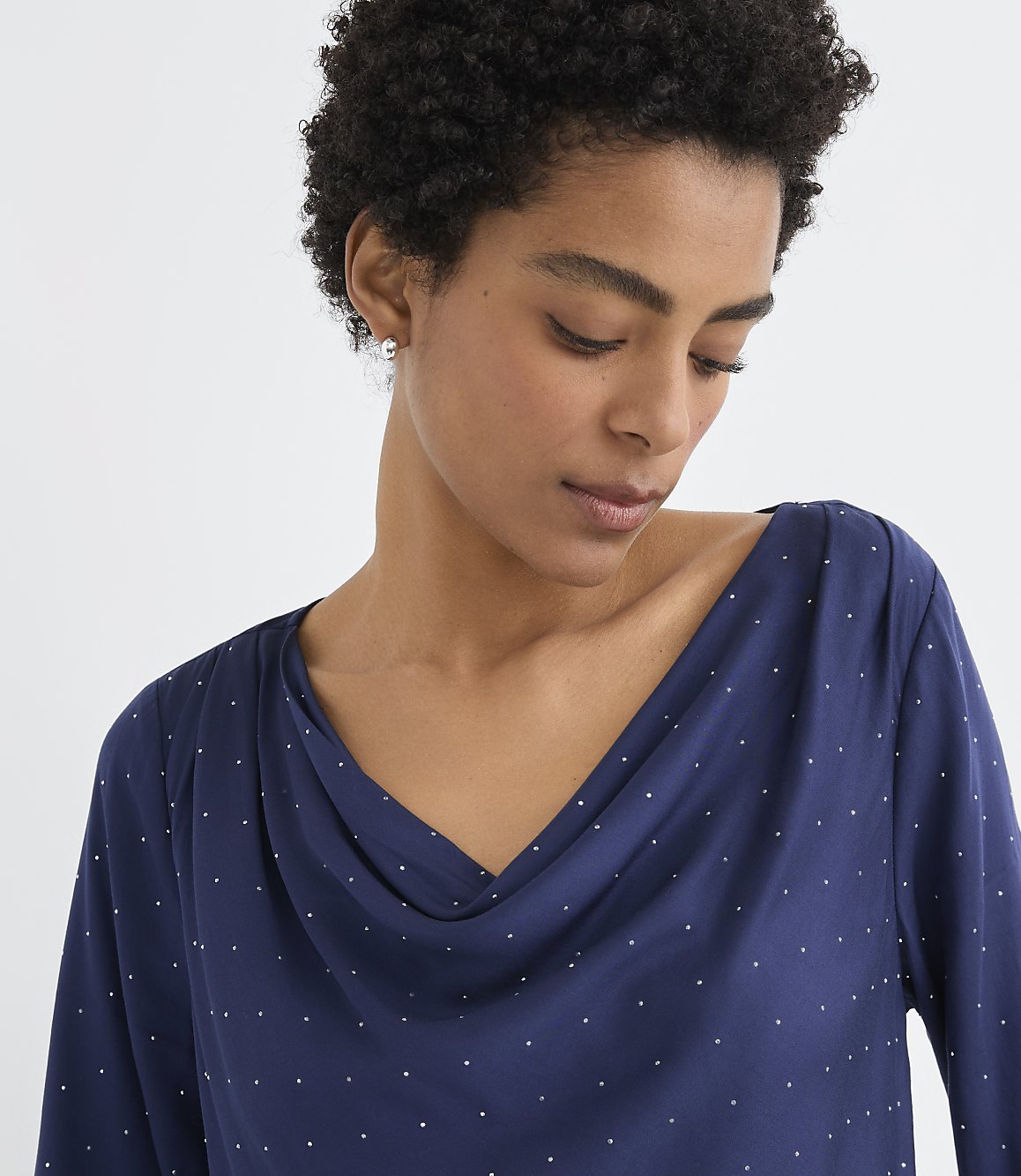 A woman with curly dark hair wearing a navy blue polka-dot top poses against a plain white background.