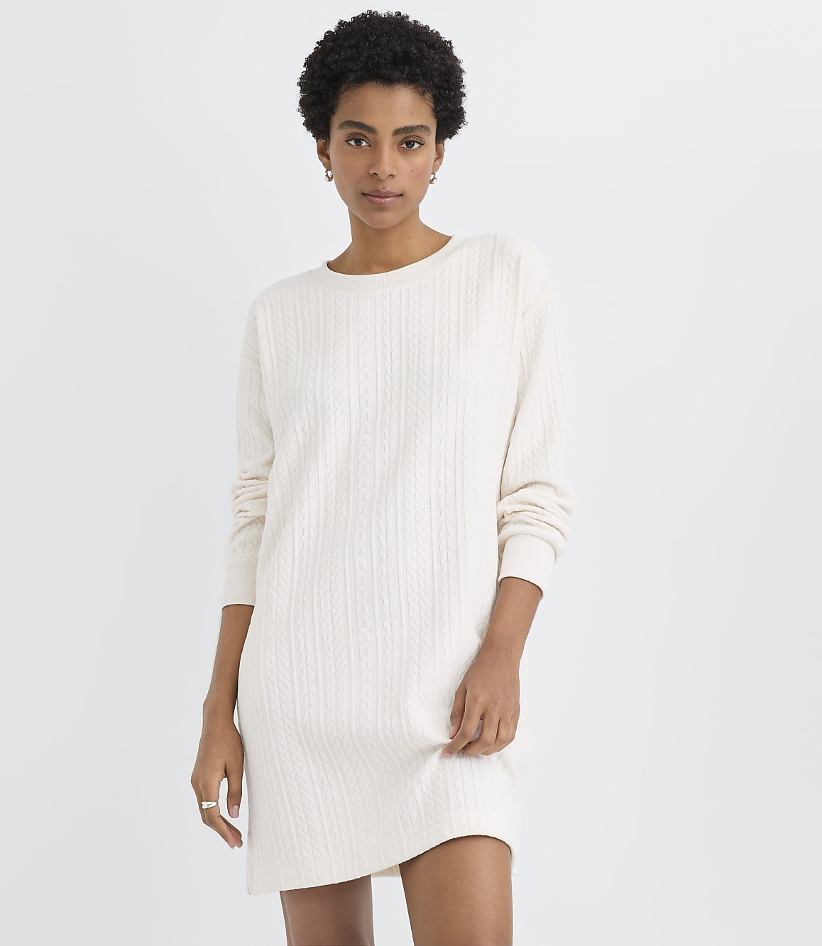 A woman wearing a white cable-knit sweater dress stands against a plain, light-colored background.
