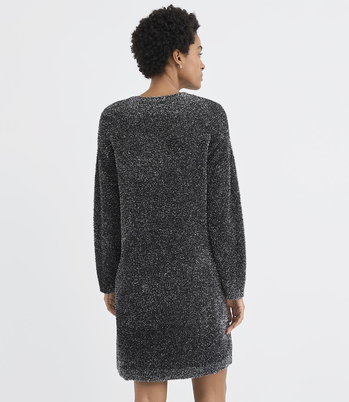 A woman with curly hair stands with her back to the camera wearing a shimmering dark grey knit dress against a plain white background.