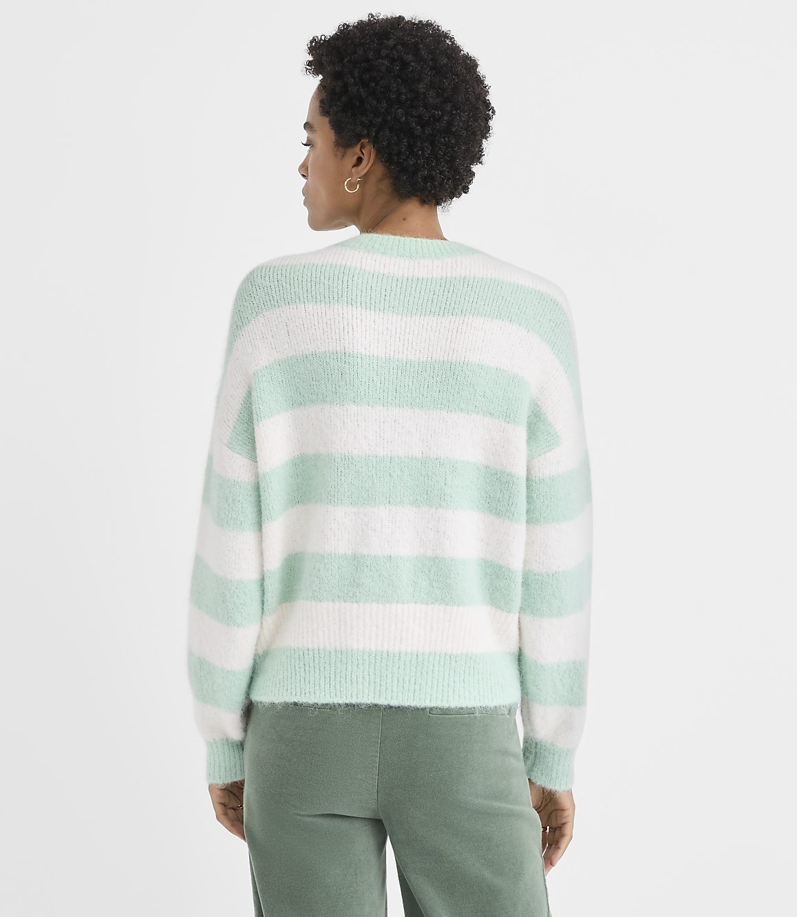 A woman with curly hair wearing a mint green and white striped sweater and green pants stands against a plain white background.