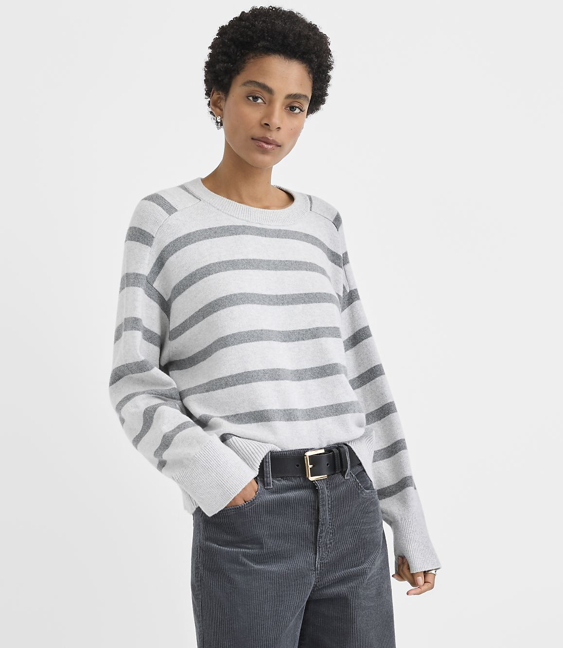 A woman wearing a grey and white striped sweater and dark grey corduroy pants stands against a plain white background.