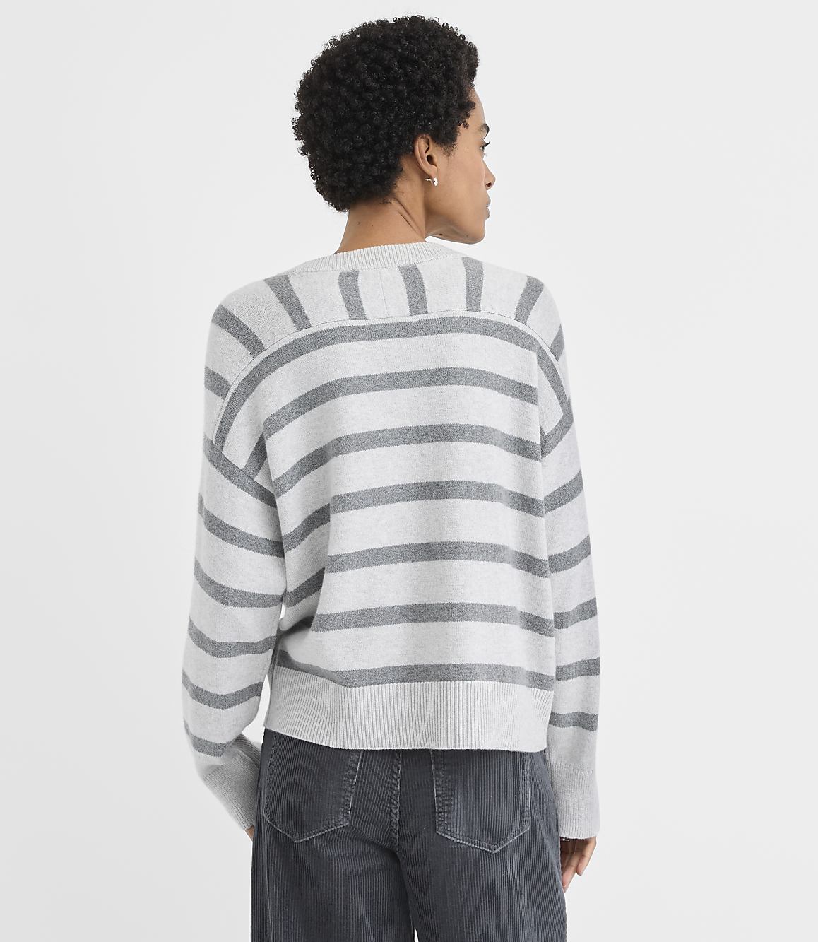 A woman wearing a grey and white striped sweater and dark grey pants stands against a plain white background.