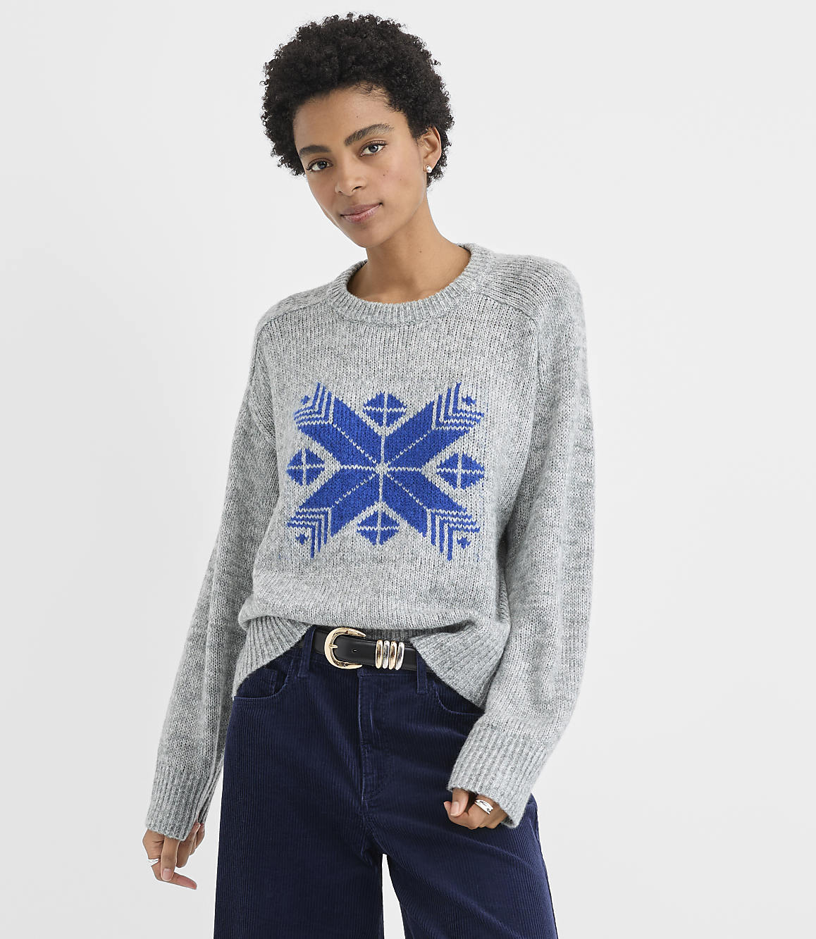 A woman wearing a grey sweater with a blue pattern and navy blue corduroy pants stands against a plain white background.
